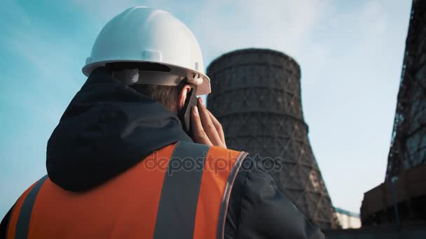 Profesní inženýr v bílou helmu a bunda se dívá na tovární potrubí. Zavolá na telefonu. Z chladících věží je bílá pára nebo kouř. — Stock video