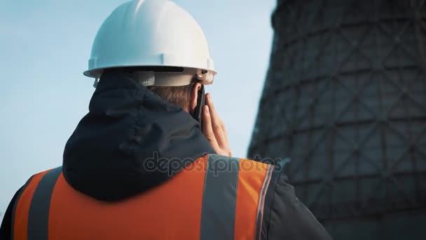 Profesní inženýr v bílou helmu a bunda se dívá na tovární potrubí. Zavolá na telefonu. Z chladících věží je bílá pára nebo kouř. — Stock video