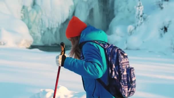 Una joven escaladora y una excursionista. Está caminando en la nieve con bastones. En el fondo, una cascada helada, témpanos de hielo y carámbanos cuelgan, el agua fluye. En busca de aventuras — Vídeo de stock