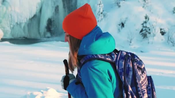 若い女の子の登山者とハイカー。杖で雪の中を歩いています。背景、凍結の滝、流氷、つららハング、に対して水の流れ。冒険を求めて — ストック動画