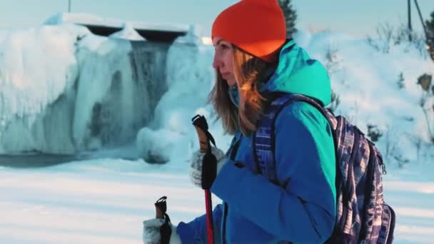 Una giovane arrampicatrice e un'escursionista. Cammina nella neve con i bastoni da passeggio. Sullo sfondo, una cascata ghiacciata, banchi di ghiaccio e ghiaccioli pendono, l'acqua scorre. Alla ricerca di avventure — Video Stock