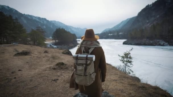 Uma jovem viajante se aproxima da beira da costa do penhasco. Ele olha para a distância para o rio gelado no inverno. Um chapéu e uma mochila de viajantes são colocados — Vídeo de Stock
