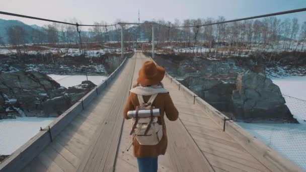 따라 A 젊은 여자 여행자 얼어붙은 겨울 강 다리를 걷는다. 모자와 여행자 배낭 넣어. — 비디오