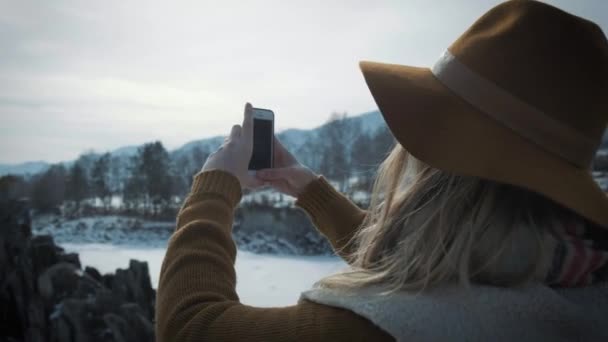 Een jong meisje reiziger neemt een foto op een smartphone. Foto's een bevroren winter rivier. Een hoed en een rugzak van reizigers zijn zetten. — Stockvideo