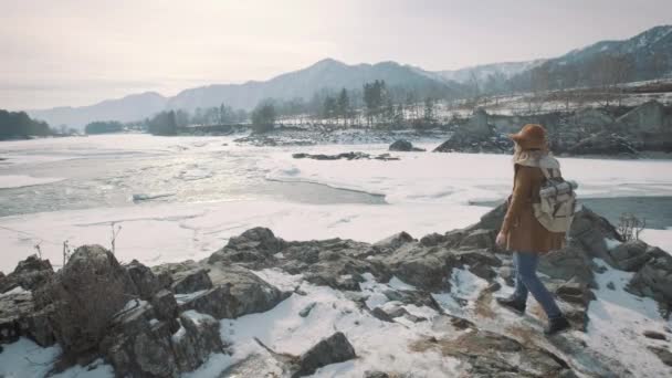 一个年轻的女孩旅行者接近悬崖岸边的边缘。冬天, 他望向远处的冰冻河。一顶帽子和一个旅行者背包放在 — 图库视频影像