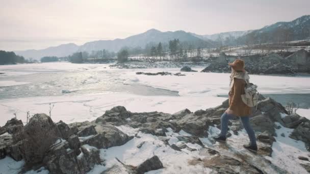 Bir genç kız gezgin bir smartphone bir fotoğrafı çeker. Donmuş kış nehir fotoğraflar. Bir şapka ve bir seyahat etmek sırt çantası koymak. — Stok video