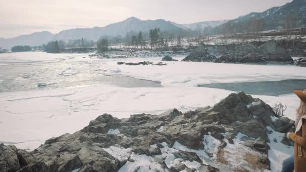 Une jeune voyageuse prend une photo sur un smartphone. Photographie une rivière gelée d'hiver. Un chapeau et un sac à dos de voyageurs sont enfilés . — Video