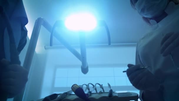 Два врача стоматолога в маске улыбаются, держа рот зеркало и крюк, включает свет стоматологической лампы, огни в рот пациента, стоя на пациенте, глядя на камеру, пытаясь — стоковое видео