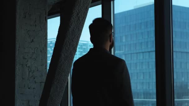 Молодой успешный бизнесмен смотрит в окно на центр города. Высокое бизнес-здание. Офис в мансарде — стоковое видео