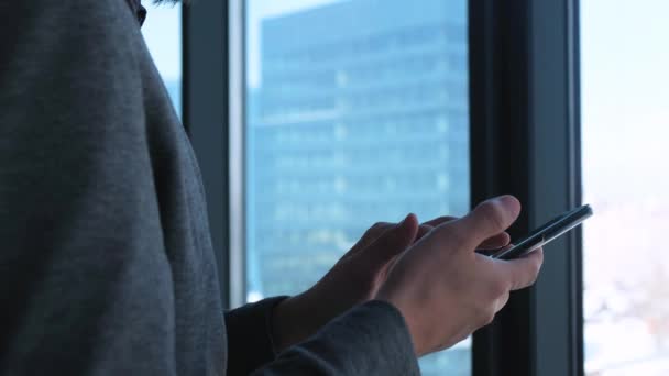 Joven empresario exitoso con barba hace una llamada en un teléfono inteligente en un fondo de ventana con una vista del centro de la ciudad. Edificio de negocios alto. Oficina en estilo loft . — Vídeos de Stock