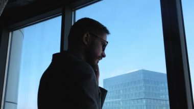 Sakallı genç işadamı smartphone cep telefonu ile şehir merkezine bakan bir pencere arka plan üzerinde bir çağrı yapar. Bir hatasının işinde. Yüksek işletme binası