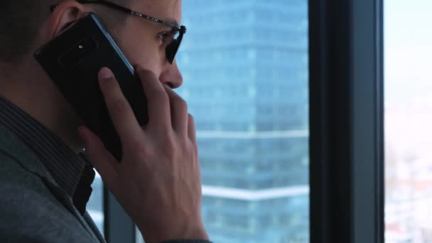 Joven empresario exitoso con barba hace una llamada en un teléfono inteligente en un fondo de ventana con una vista del centro de la ciudad. Edificio de negocios alto. Oficina en estilo loft . — Vídeos de Stock