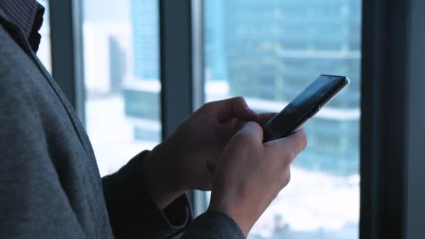 Mladý úspěšný podnikatel s bradkou zavolá na smartphone na pozadí okna s výhledem na centrum města. Vysoké obchodní budovy. Úřad v loft stylu. — Stock video