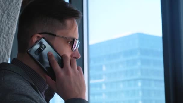 Joven empresario exitoso con barba hace una llamada en un teléfono inteligente en un fondo de ventana con una vista del centro de la ciudad. Edificio de negocios alto. Oficina en estilo loft . — Vídeos de Stock