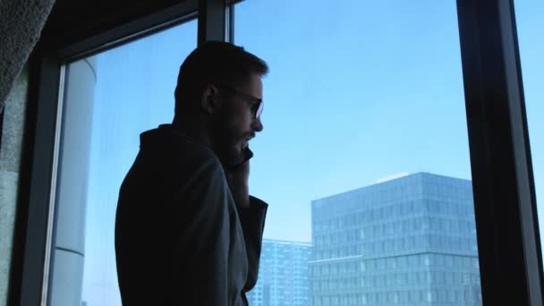 Jovem empresário com barba faz uma chamada em um smartphone no fundo de uma janela com vista para o centro da cidade. Está experimentando um fracasso no negócio. Edifício empresarial elevado — Vídeo de Stock