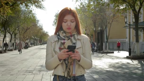 Молодая девушка стоит на городской улице. Держать смартфон, набирать текстовое сообщение. Общался с друзьями в социальных сетях. Снаружи — стоковое видео