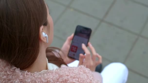 Belle jeune fille écoute de la musique grâce à des écouteurs blancs sans fil. Assis sur un banc dans la rue à l'automne. Il tient un smartphone dans sa main, change la chanson — Video