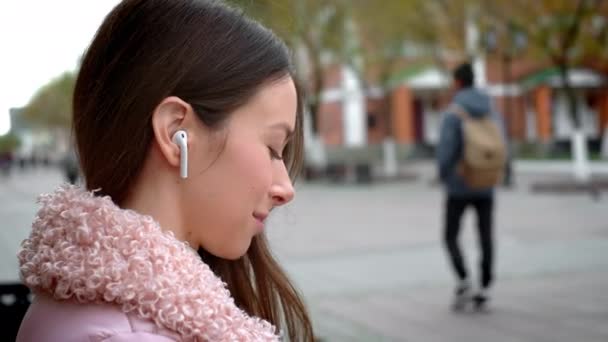 Bella ragazza sta ascoltando musica attraverso cuffie bianche senza fili. Si siede su una panchina in strada in autunno. Tiene uno smartphone in mano, cambia la canzone — Video Stock