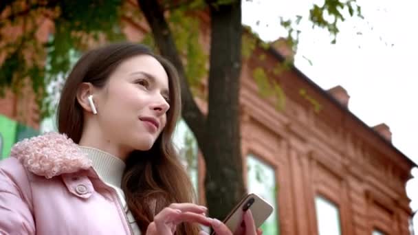 Милая молодая девушка слушает музыку через беспроводные белые наушники. Сидит на скамейке на улице осенью. Держит в руке смартфон, переключает песню — стоковое видео