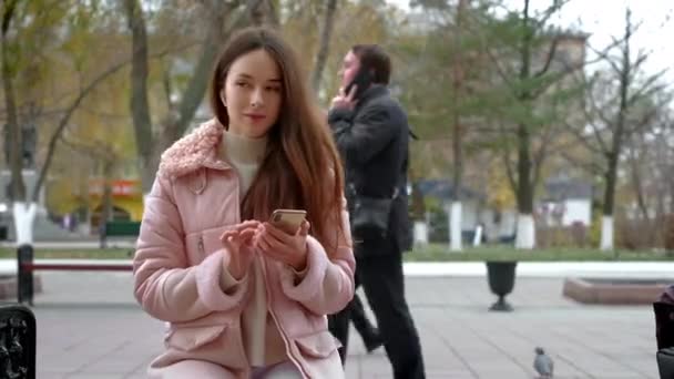 Chica joven encantadora está escuchando música a través de auriculares blancos inalámbricos. Se sienta en un banco en la calle en el otoño. Sostiene un smartphone en su mano, cambia la canción — Vídeo de stock