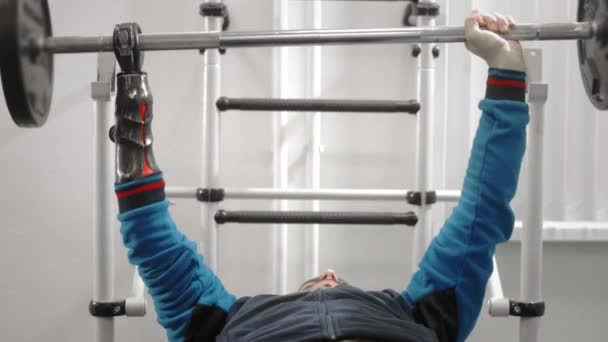 Un atleta sin mano levanta la barra mientras está acostado. Mantiene la barra con una prótesis de mano deportiva profesional. Gimnasio para Paralímpicos — Vídeo de stock