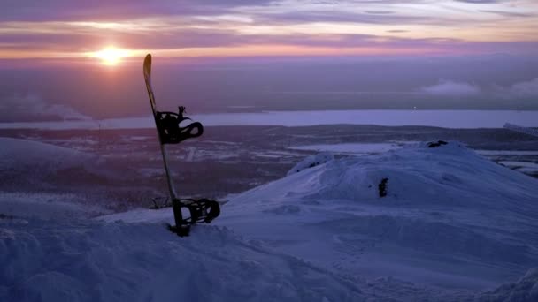 Snowboard stoi w śniegu. Utknął w zaspie śnieżnej na tle zachodu słońca. Zimowe góry pokryte śniegiem na północy Rosji, Khibiny. Krajobraz wieczorny poza kołem podbiegunowym — Wideo stockowe