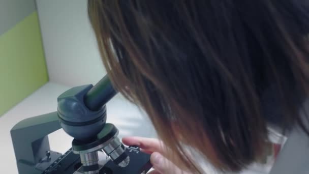 Fiatal lány tudós, mikroszkóppal nézi. Tanulmányozza a mintákat. Egy fiatal tudós tudományos kutatást folytat. Egy vírust kutat, egy járvány elleni vakcina ellenszerét keresi. — Stock videók
