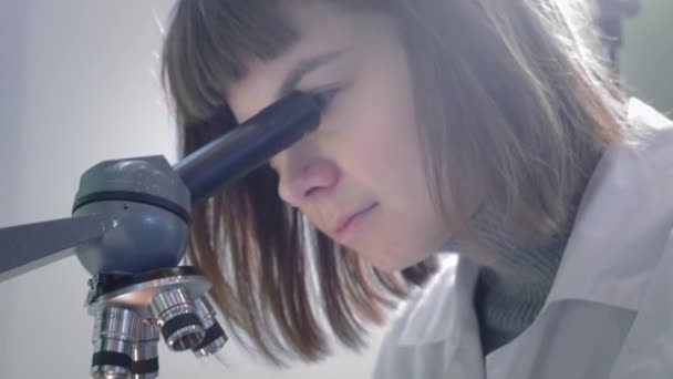 Jovem cientista olhando através de um microscópio. Ela estuda as amostras. Um jovem cientista realiza pesquisas científicas. Ela está pesquisando um vírus, procurando uma cura para uma vacina epidêmica — Vídeo de Stock