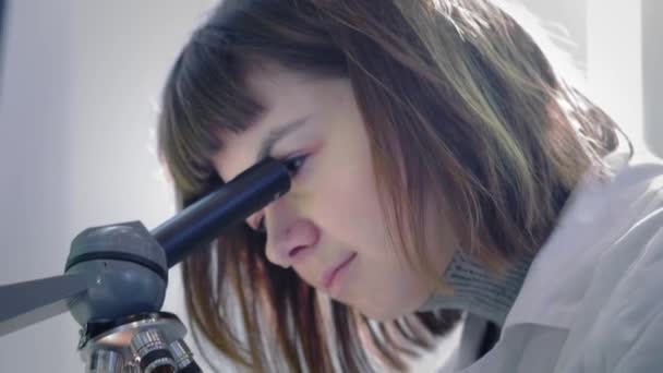 Una joven científica mirando a través de un microscopio. Estudia las muestras. Un joven científico lleva a cabo investigaciones científicas. Ella está investigando un virus, buscando una cura para una vacuna epidémica — Vídeos de Stock