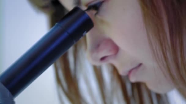 Una joven científica mirando a través de un microscopio. Estudia las muestras. Un joven científico lleva a cabo investigaciones científicas. Ella está investigando un virus, buscando una cura para una vacuna epidémica — Vídeos de Stock