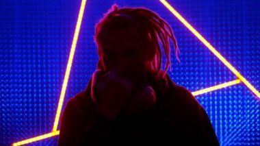 Neon ışıklı rastalı genç adam modeli. Bir kulüp stüdyosundaki neon üçgen. Dubstep ya da punk tarzı. Kirli havadan dolayı gaz maskesi takıyor. Halka açık bir yerde koronavirüsten korunuyor.