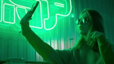 Güneş gözlüklü şık kız blogcu akıllı telefon kullanarak selfie ya da video çekiyor. Çekici esmer genç kadın kendine cep telefonuyla bakıyor ve kısa saçlarına dokunuyor. Yeşil neon ışığı