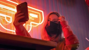 Genç, güzel, modaya uygun bir kız siyah tıbbi maskeli bir selfie çekiyor. Neon ışıklarının arkasındaki bir barda ya da kulüpte. Çin koronavirüsü salgınına karşı korur. Gezegende salgın var.