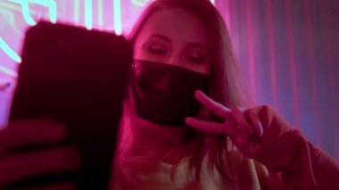 Genç, güzel, modaya uygun bir kız siyah tıbbi maskeli bir selfie çekiyor. Neon ışıklarının arkasındaki bir barda ya da kulüpte. Çin koronavirüsü salgınına karşı korur. Gezegende salgın var.