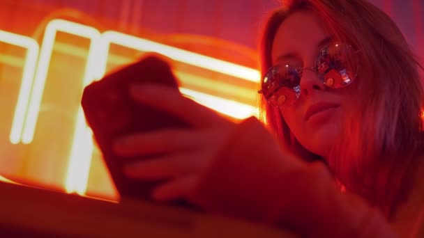 Portrait of attractive smiling girl using online social media and listening music. Cute young woman typing message on smartphone and smile. Dressed casual street style. Colorful red neon lights. — Stock Video