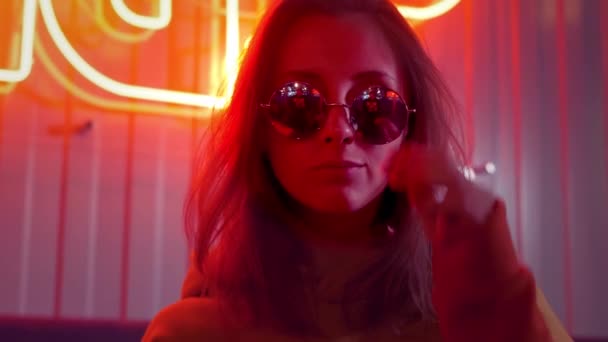 Young stylish beautiful girl model in sunglasses dancing in a bar or club. Red neon sign on the wall. Neon red lights in a grunge setting. Playful movements, flirts with the camera — Stock Video