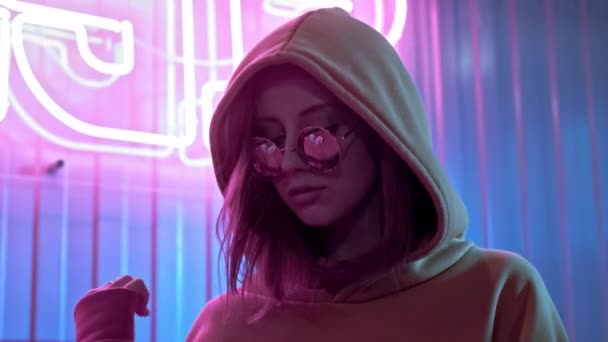 Young stylish beautiful girl model in sunglasses dancing in a bar or club. Red neon sign on the wall. Neon pink lights in a grunge setting. Playful movements, flirts with the camera — Stock Video