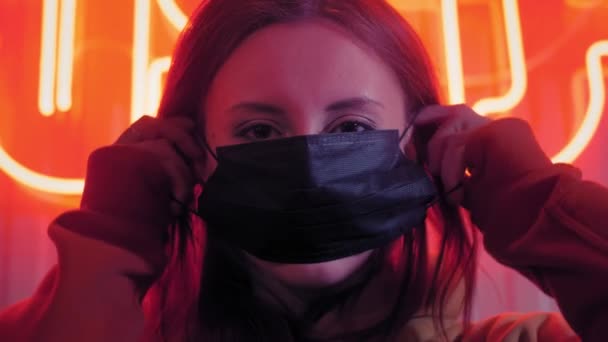 Mladá krásná módní dívka si nasadí černou lékařskou masku. V baru nebo klubu na pozadí neonových světel. Chrání před epidemiemi koronaviru. Pandemie na planetě. — Stock video