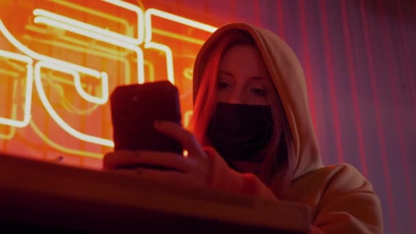 Young beautiful fashionable girl uses a smartphone in a black medical mask. In a bar or club on background of neon lights. Protects against the epidemic of Chinese coronavirus. Pandemic on the planet — Stock Video