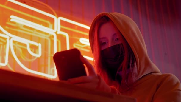 Ung vacker fashionabla flicka använder en smartphone i en svart medicinsk mask. I en bar eller klubb på bakgrund av neonljus. Skyddar mot epidemin av kinesisk coronavirus. Pandemi på planeten — Stockvideo