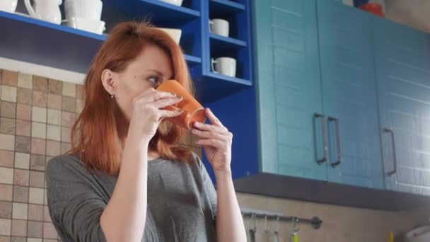 Attractive girl with red hair drinks morning coffee in the home kitchen. Freelancer morning. Enjoys a sleepy morning in the kitchen. — Stock Video