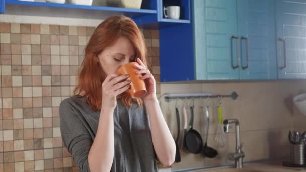 Kızıl saçlı çekici kız sabah kahvesini ev mutfağında içiyor. Freelancer sabahı. Mutfakta uykulu bir sabahın tadını çıkarıyor.. — Stok video
