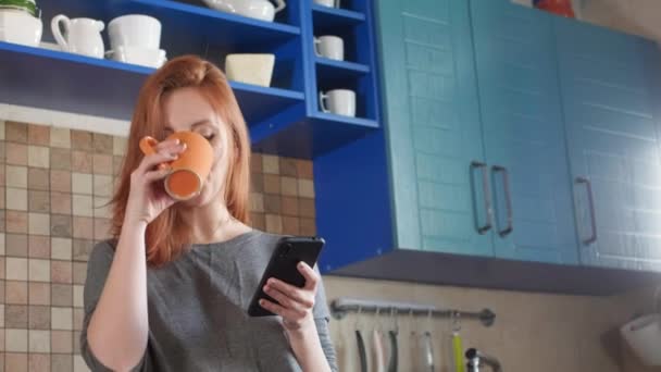 Attraente ragazza con i capelli rossi beve caffè mattutino nella cucina di casa. Mattina da freelance. Utilizza uno smartphone, guarda un notiziario o un social network. Scrive un messaggio al messaggero — Video Stock