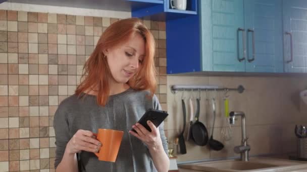 Chica atractiva con el pelo rojo bebe café de la mañana en la cocina del hogar. Hablando en un smartphone. Hace una llamada matutina a un amigo o pide comida a domicilio . — Vídeo de stock