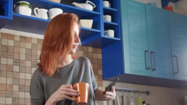 빨간 머리의 매력적 인 여자가 아침 커피를 집 부엌에서 마십니다. 스마트폰으로 얘기하는 거죠. 친구에게 아침에 전화를 걸 거나 음식을 배달하라는 지시를 한다. — 비디오