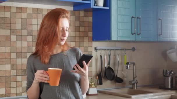 Jolie fille aux cheveux roux boit le café du matin dans la cuisine de la maison. Parler sur un smartphone. Prend un appel matinal à un ami ou commande la livraison de nourriture . — Video