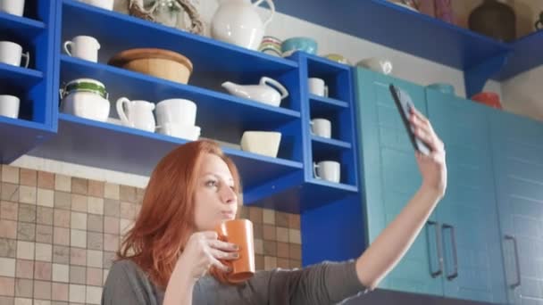 Приваблива дівчина з рудим волоссям п'є ранкову каву на домашній кухні. Робить селфі на смартфоні для соціальних мереж. Красива жінка використовує телефон для селфі фото на сучасній кухні . — стокове відео