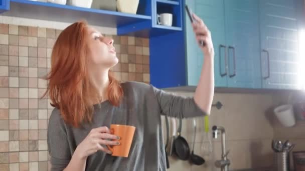 Menina atraente com bebidas de cabelo vermelho café da manhã na cozinha da casa. Faz uma selfie em um smartphone para redes sociais. Mulher bonita usando telefone para foto selfie na cozinha moderna . — Vídeo de Stock