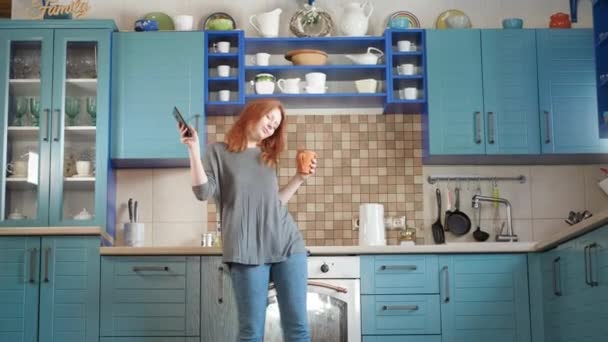 Танцевать весело, слушать музыку на смартфоне. Привлекательная девушка с рыжими волосами пьет утренний кофе на домашней кухне. Молодая девушка танцует и пользуется мобильным телефоном. Рыжая женщина танцует на кухне — стоковое видео