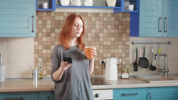 跳舞有趣，在智能手机上听音乐。迷人的红头发女孩在自家厨房喝早茶。年轻的女性跳舞和使用手机。红头发女人在厨房里跳舞 — 图库视频影像